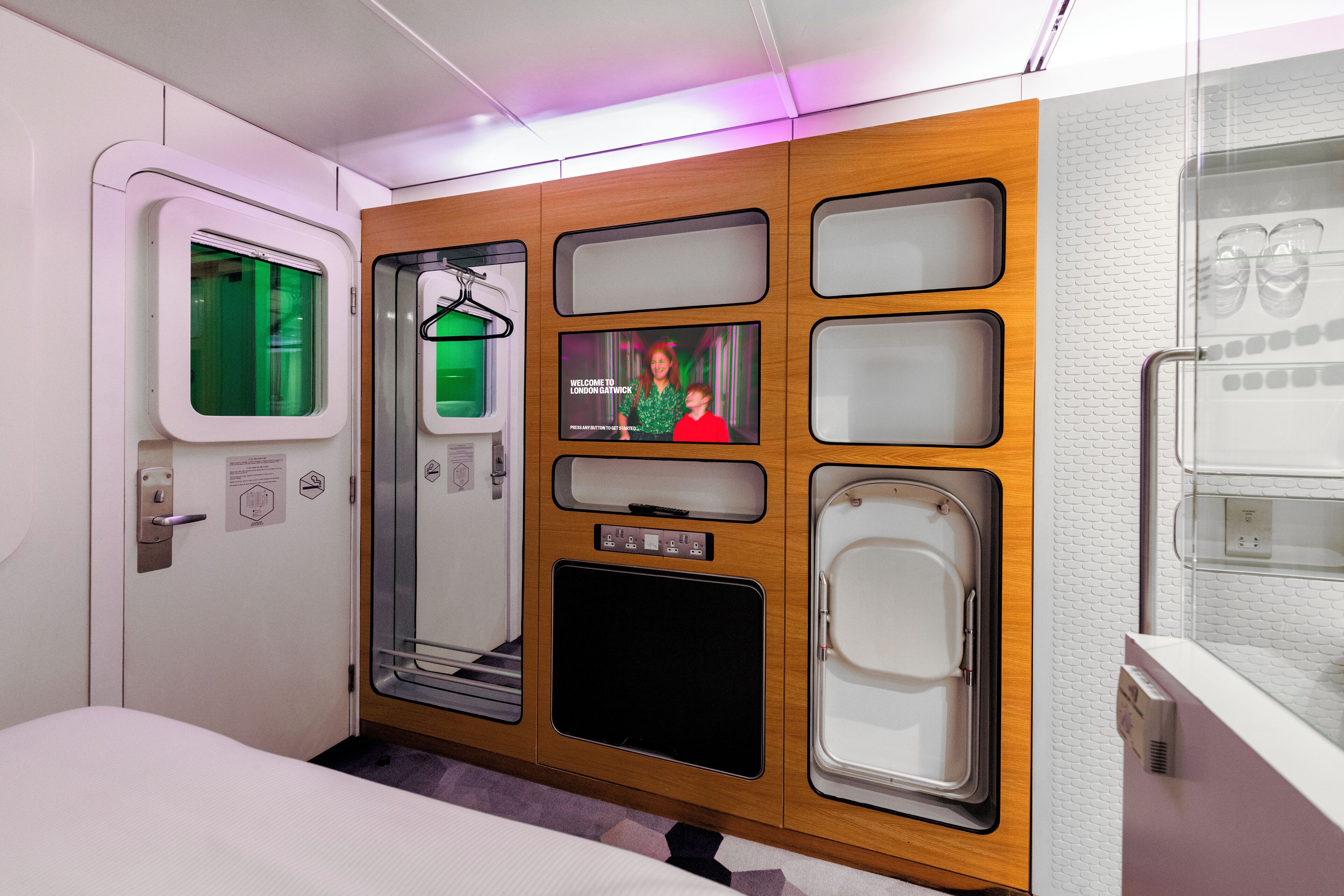 Un compartiment de train moderne avec un mur en panneaux de bois, un écran de télévision affichant un message de bienvenue et une porte en verre dotée d’une poignée.