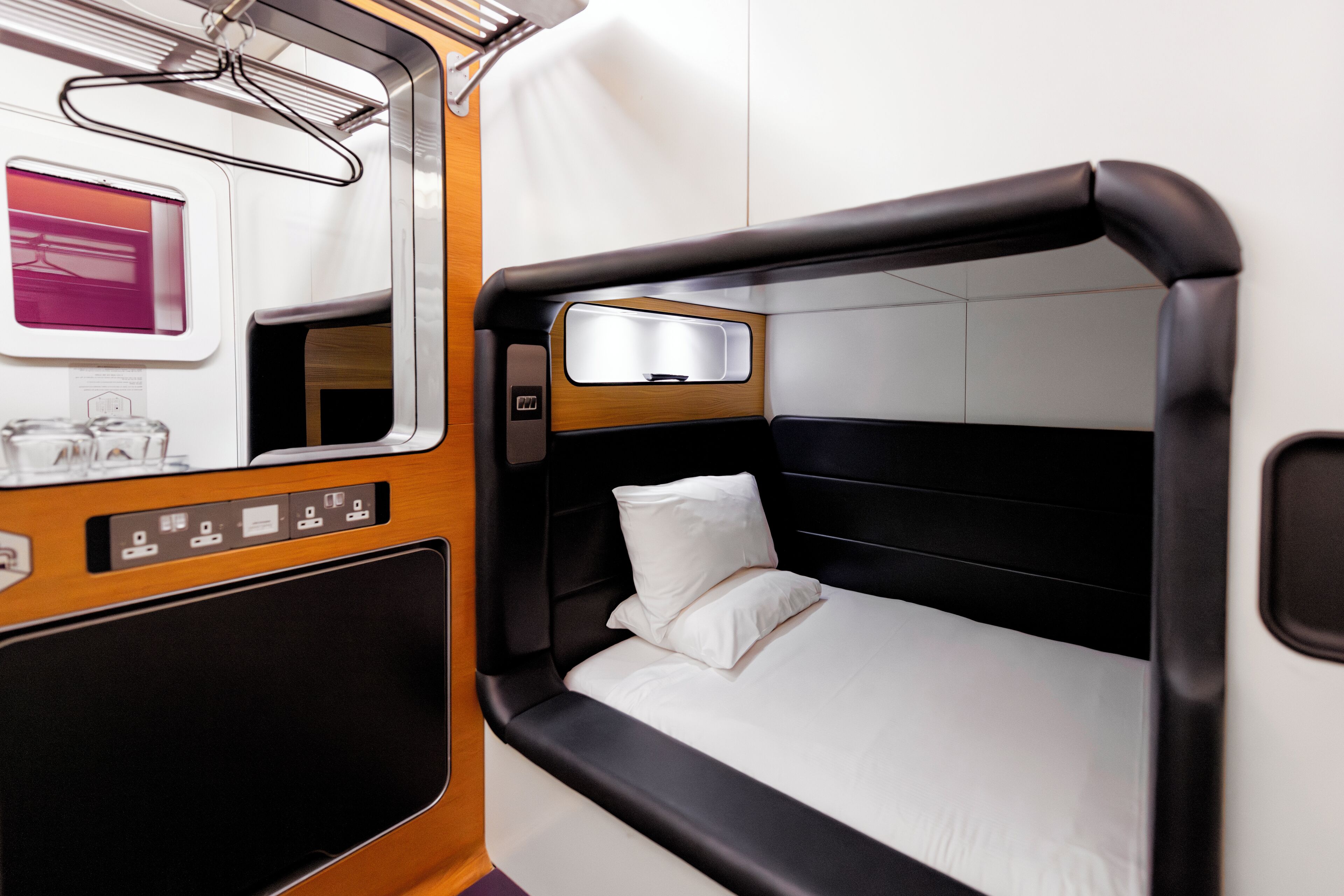 A compact, modern train cabin with a single bed, a small table with a mirror, and a window with a view of the outside.