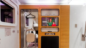 A compact, well-lit room with a bed, a television screen displaying a welcome message, a small seating area, and a door with a window. - YotelAIR London Gatwick (Gatwick)
