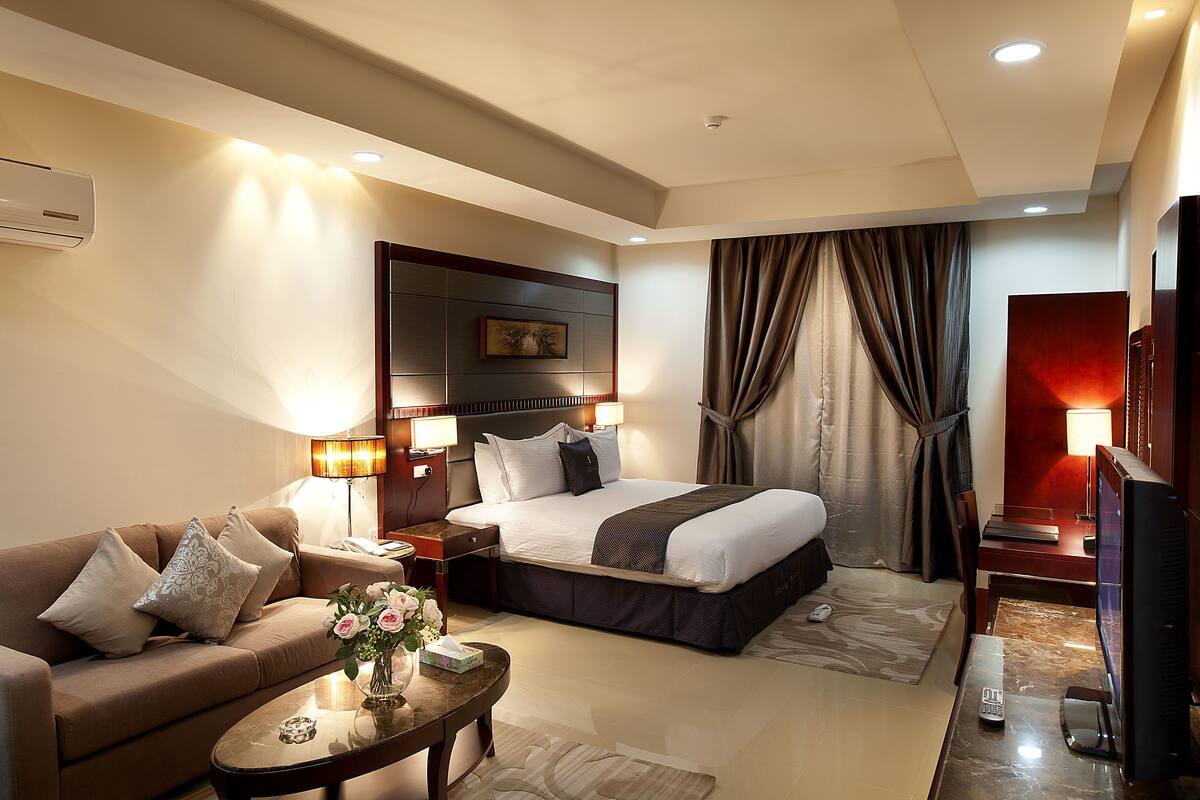 A hotel room with a bed, a sofa, a TV, and a desk with a lamp.