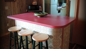 Una cocina con una isla rosa, taburetes verdes, paredes de madera y un microondas.
