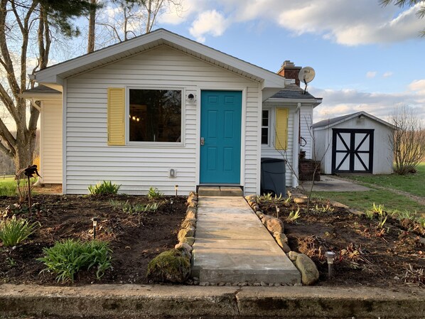 Una casa de un piso con puerta azul, una antena parabólica y un cobertizo pequeño al fondo.