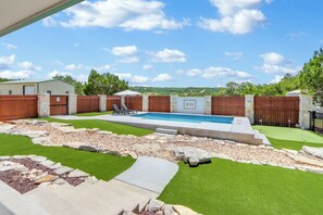 A backyard with a swimming pool, artificial grass, stone pathways, and a wooden fence. - Artista I 2/2 with New Pool, Hot tub,  Pool Table (Wimberley)