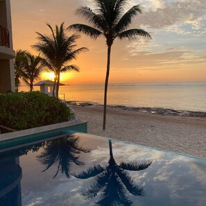 Piscina com reflexo de palmeiras e pôr do sol sobre o oceano.