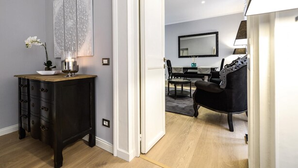 A modern living room with a dark wooden cabinet, a white vase with flowers, a black sofa, and a dining area with a table and chairs. - Belsiana Apartment - Two Bedroom Apartment, Sleeps 6 (Roma)
