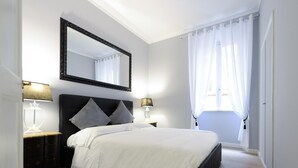 A bedroom with a large bed, a nightstand with a lamp, a dresser, a mirror, and a window with curtains. - Belsiana Apartment - Two Bedroom Apartment, Sleeps 6 (Roma)