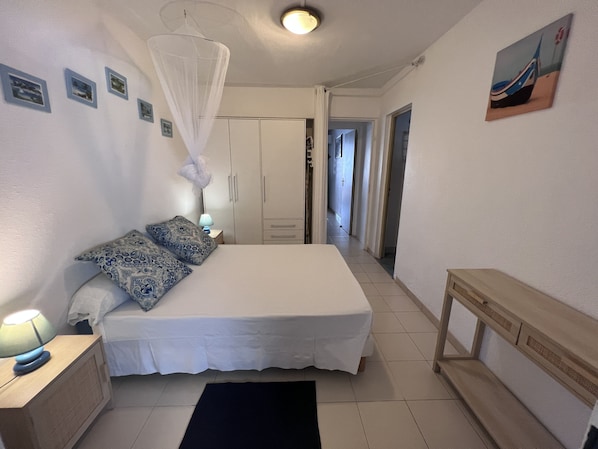 Une chambre à coucher avec un lit, une table de chevet, une lampe et un baldaquin. Il y a une porte qui mène à une autre pièce et un tableau accroché au mur.