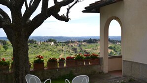 Eine Terrasse mit zwei weißen Stühlen, einem Baum und Blick auf eine grüne Landschaft mit Gebäuden in der Ferne.