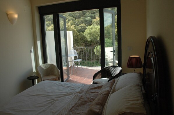 A bedroom with a bed, a chair, a small table, a lamp, and a balcony with a view of trees.