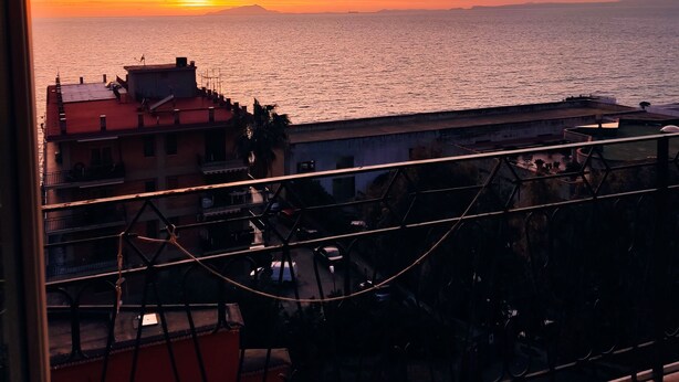 Vista del atardecer desde un balcón con vista a una masa de agua, con edificios y una valla en primer plano.