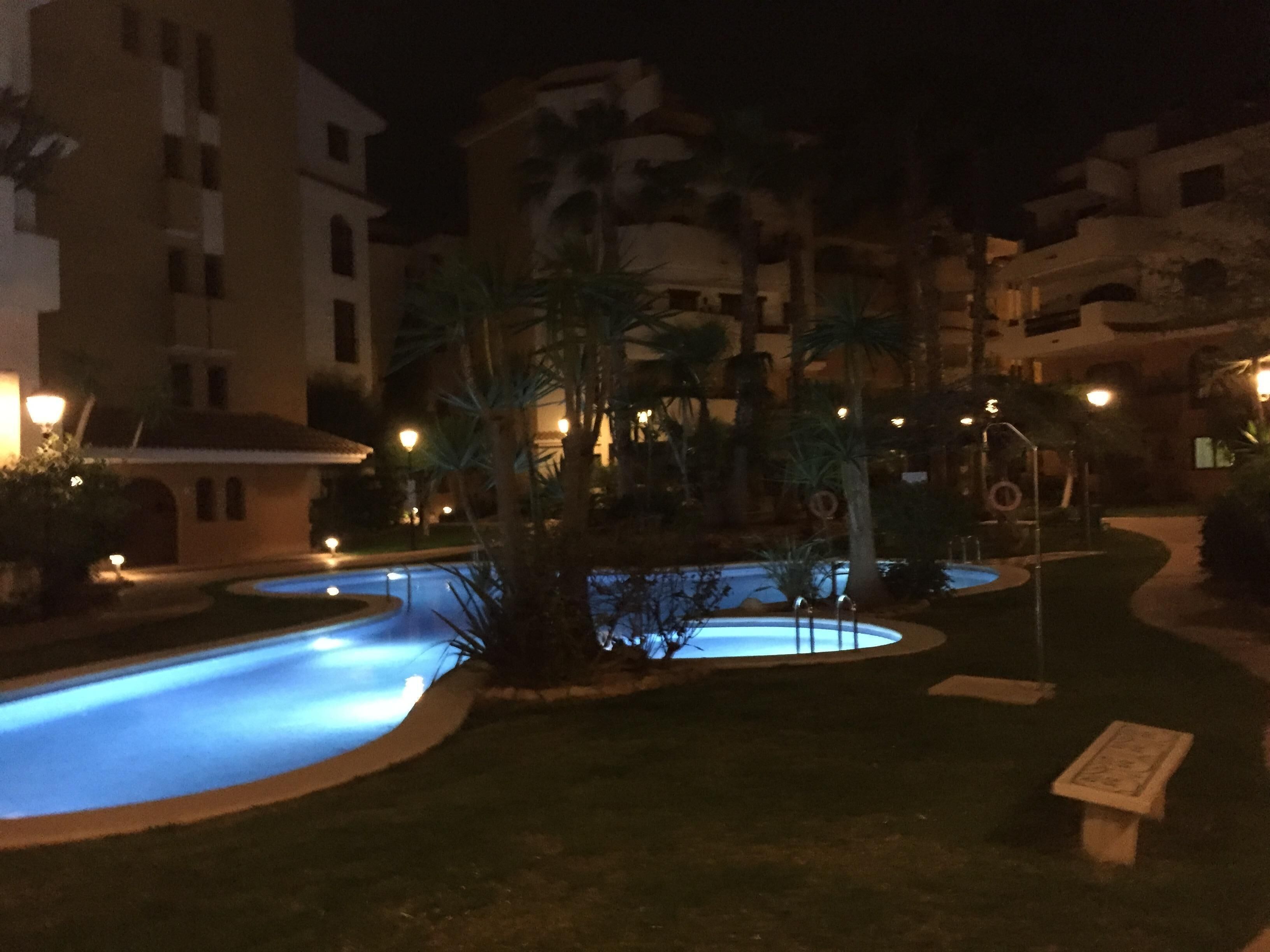 Vista nocturna de un patio de hotel con piscina, palmeras y farolas iluminadas.