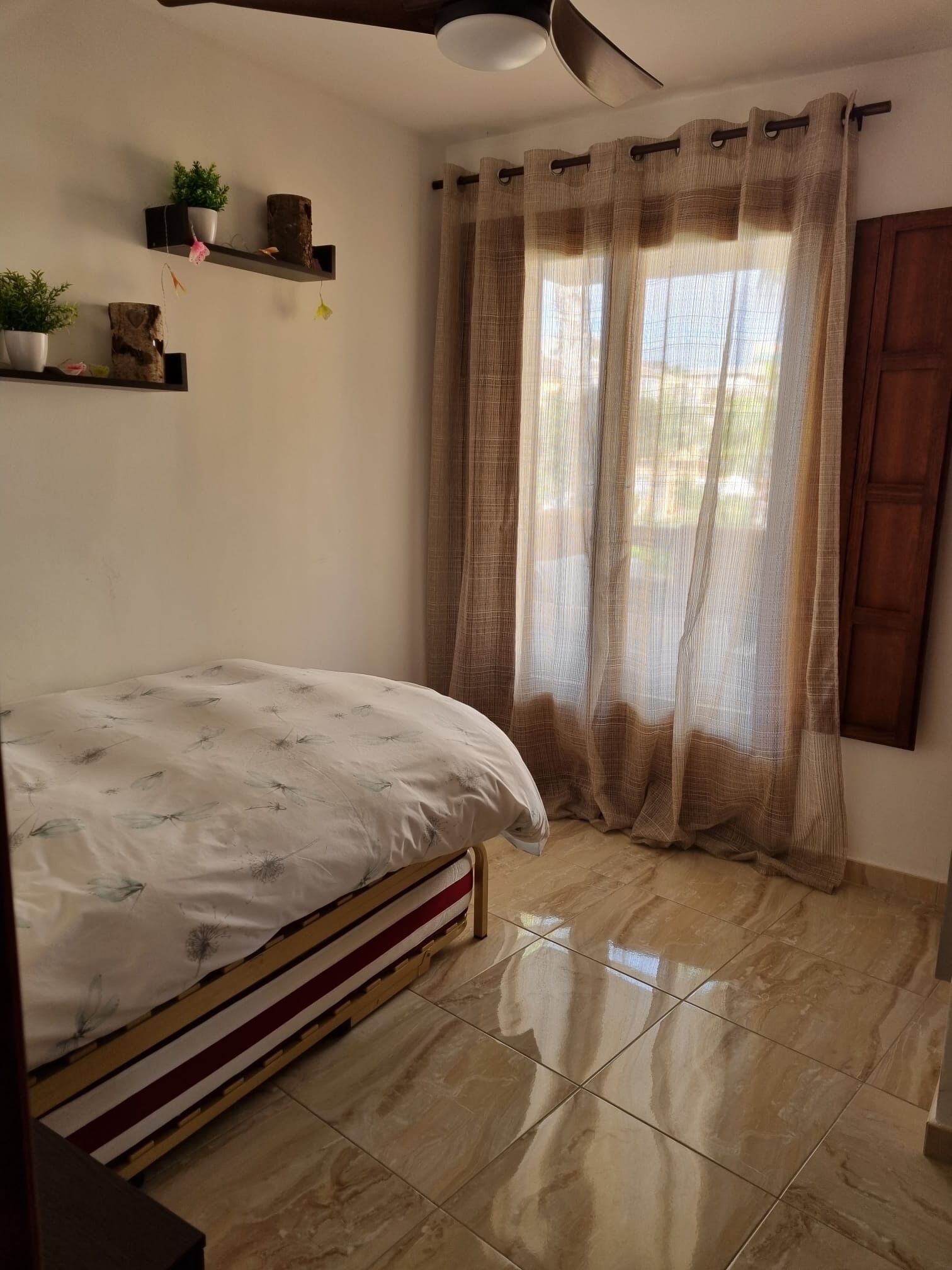 Ein Schlafzimmer mit Bett, Deckenventilator, Fenster mit Vorhängen und einer Pflanzenablage.