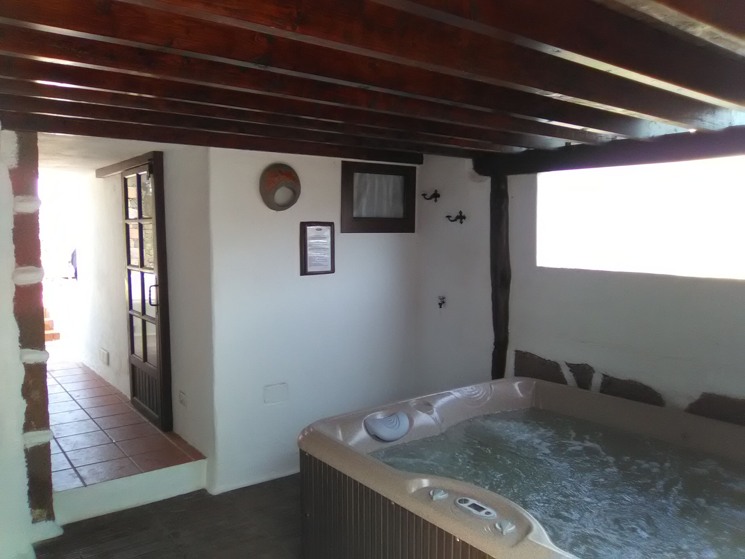A room with a hot tub, wooden beams on the ceiling, a small window, and a door leading outside.