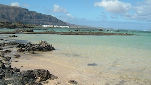 A beach with clear turquoise water, rocky shoreline, and white buildings in the background. - Casa Rural La Molina large apartment in an old motor mill from 1912 (Maguez  -Lanzarote)