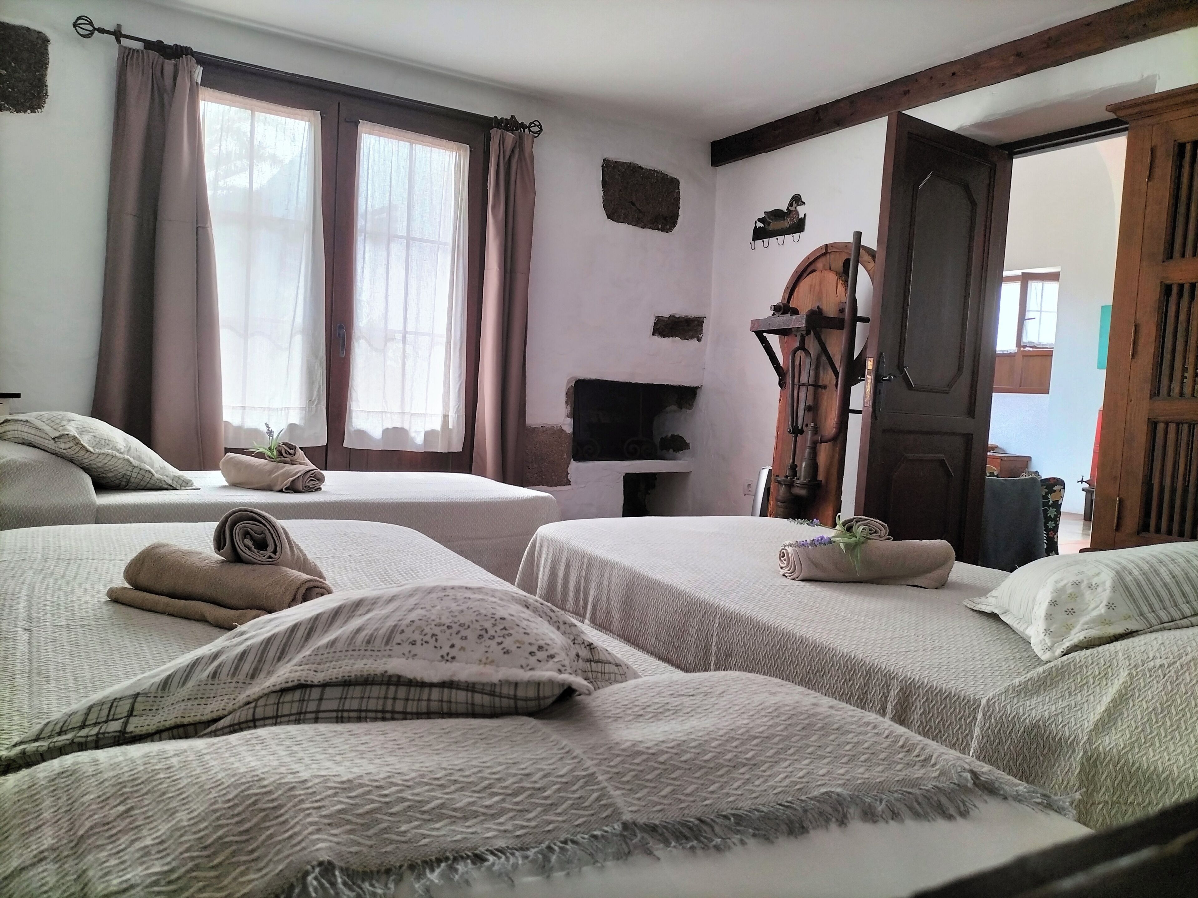 Ein Zimmer mit zwei Betten, jeweils mit ordentlich gefalteten Handtüchern und Kissen. Eine Holzbank mit rustikalem Design und ein kleines Regal mit einem dekorativen Gegenstand. Ein Fenster mit Vorhängen lässt natürliches Licht in den Raum.