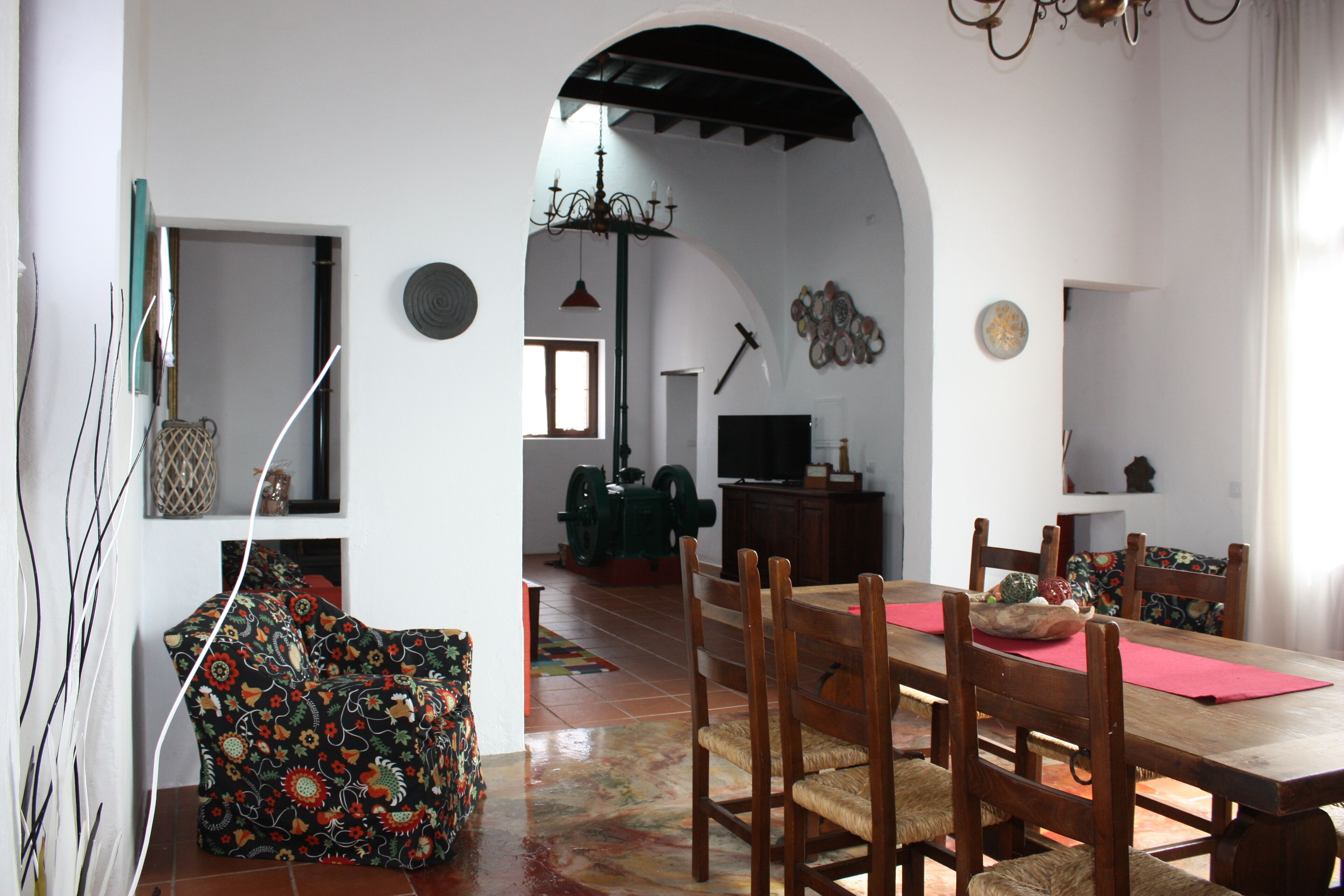 A dining area with a wooden table, chairs, and a red placemat. There is a patterned sofa and a chandelier in the background.