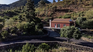 Un bâtiment rouge coiffé d’un toit de tuiles, entouré d’un mur de pierre et d’une végétation luxuriante, avec une chaîne de montagnes en arrière-plan.