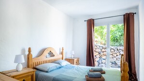 Une chambre à coucher avec un lit en bois, deux tables de chevet, une commode et une vue sur la verdure par une fenêtre ornée de rideaux.