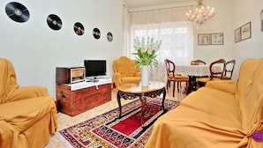 A living room with a sofa set, a wooden cabinet with a television, a table with a vase, and a chandelier.