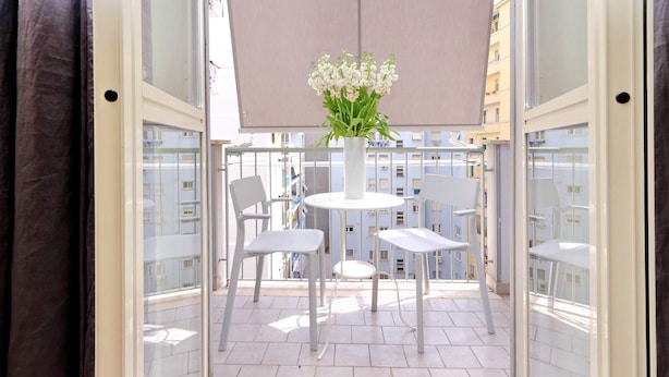 A balcony with a table and chairs, a vase with flowers, and a view of buildings.
