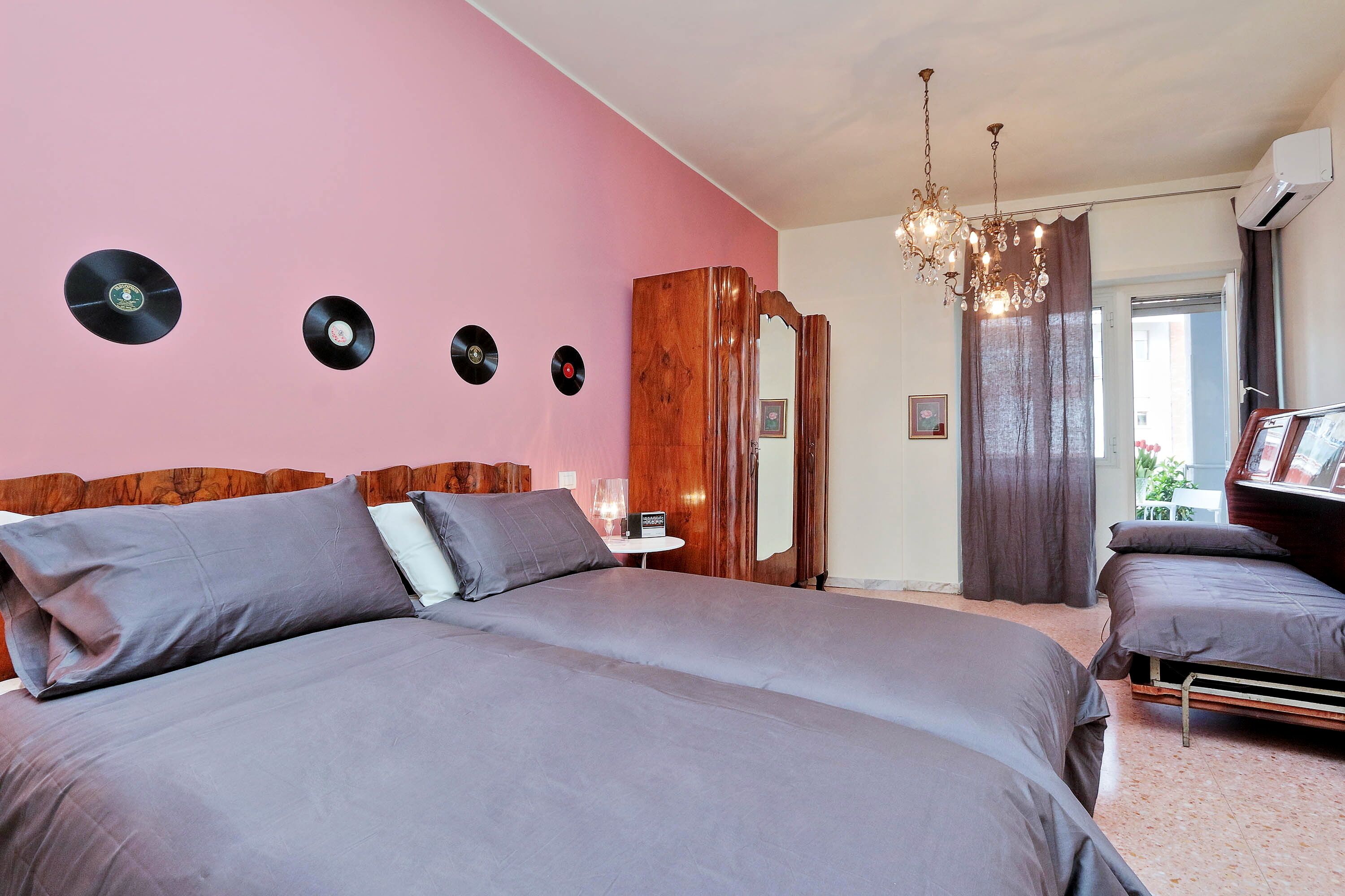 Une chambre avec un grand lit, un plus petit lit, une armoire en bois, un miroir, un lustre et une fenêtre avec des rideaux.