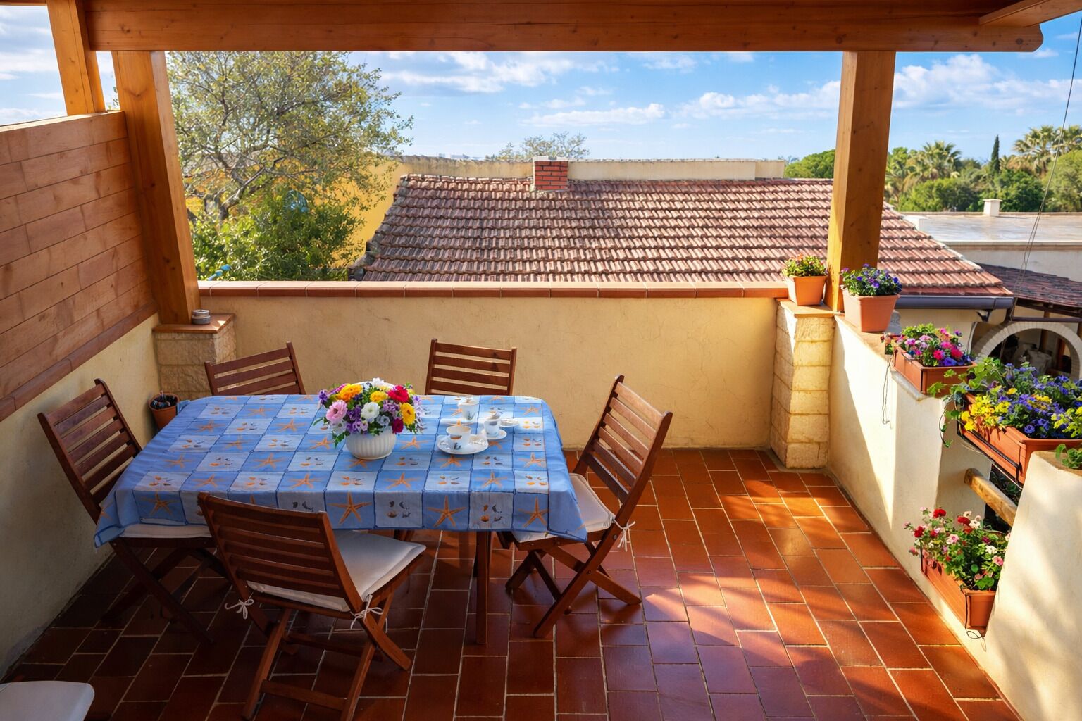 A covered patio with a table set for four, surrounded by wooden chairs, a blue tablecloth, and a floral centerpiece.