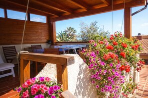 Un espace extérieur aménagé en terrasse, avec des meubles en bois, une table et un jardinier suspendu où s’épanouissent des fleurs aux couleurs vives.