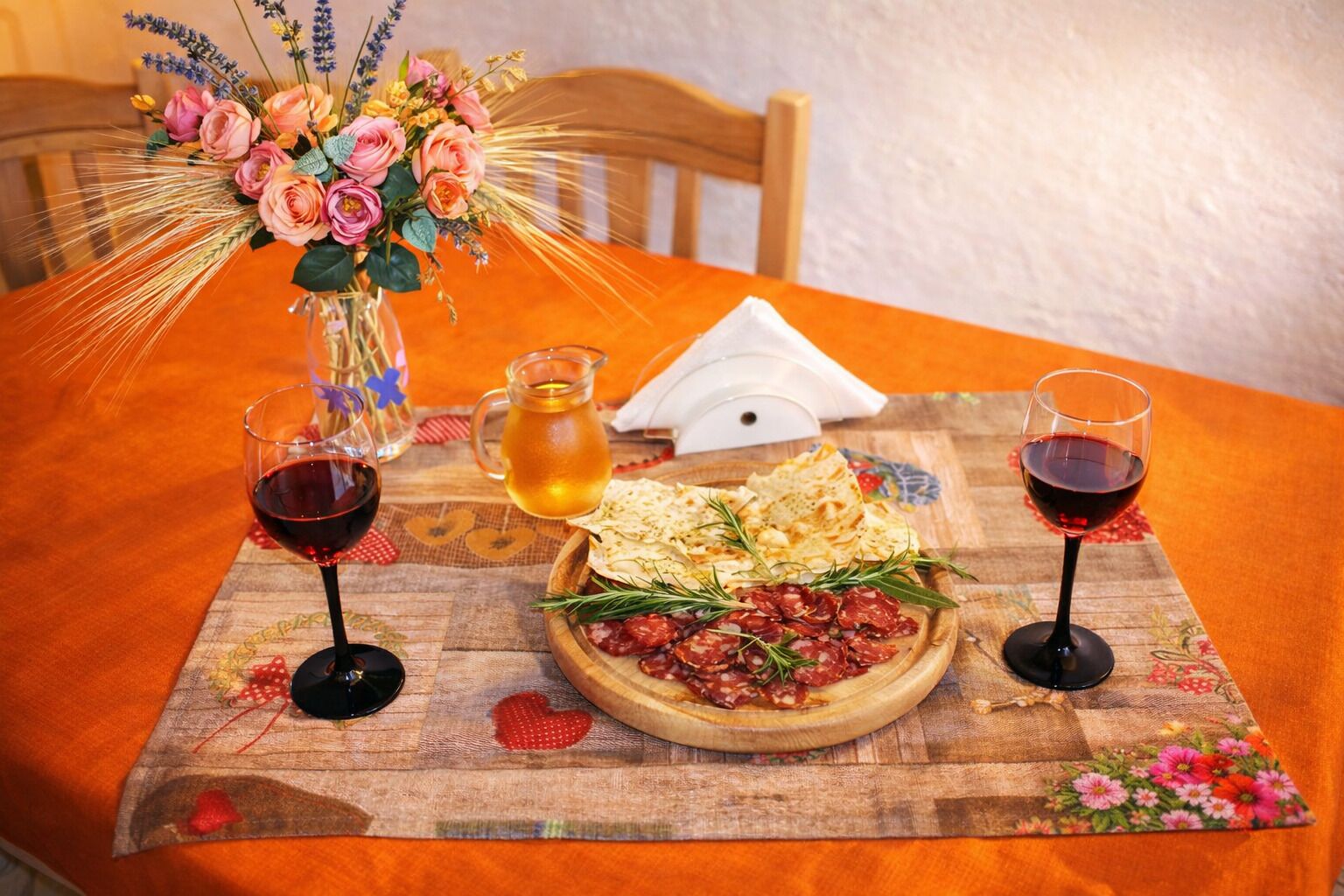 Ein Tisch ist gedeckt mit einem floralen Mittelstück, einer Kanne, einer Platte mit Wurstwaren und zwei Gläsern Rotwein.