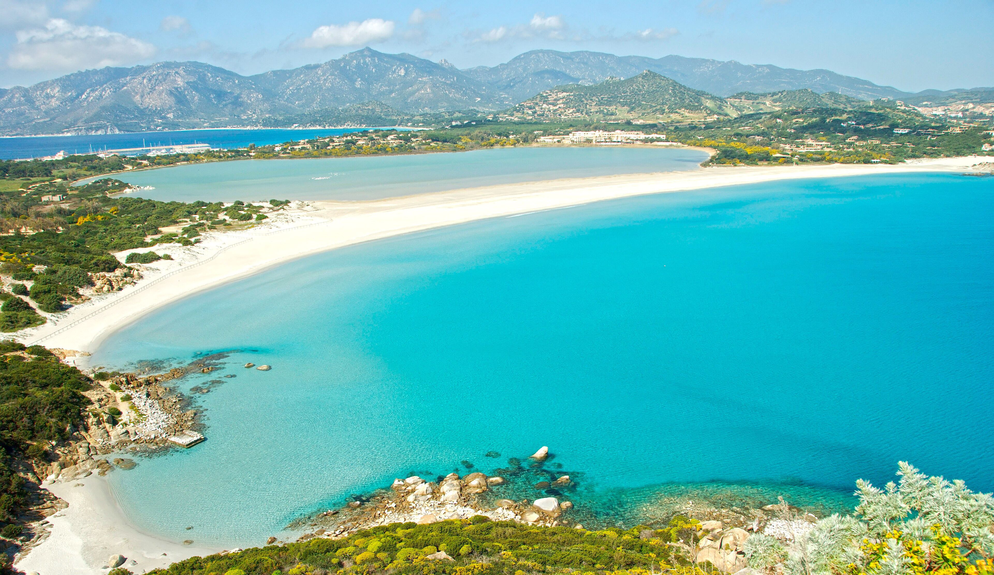 A coastal landscape with a sandy beach, clear turquoise waters, and mountainous terrain in the background.