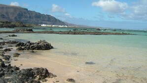 Une plage à l’eau turquoise et limpide, avec une côte rocheuse et des bâtiments blancs en arrière-plan.