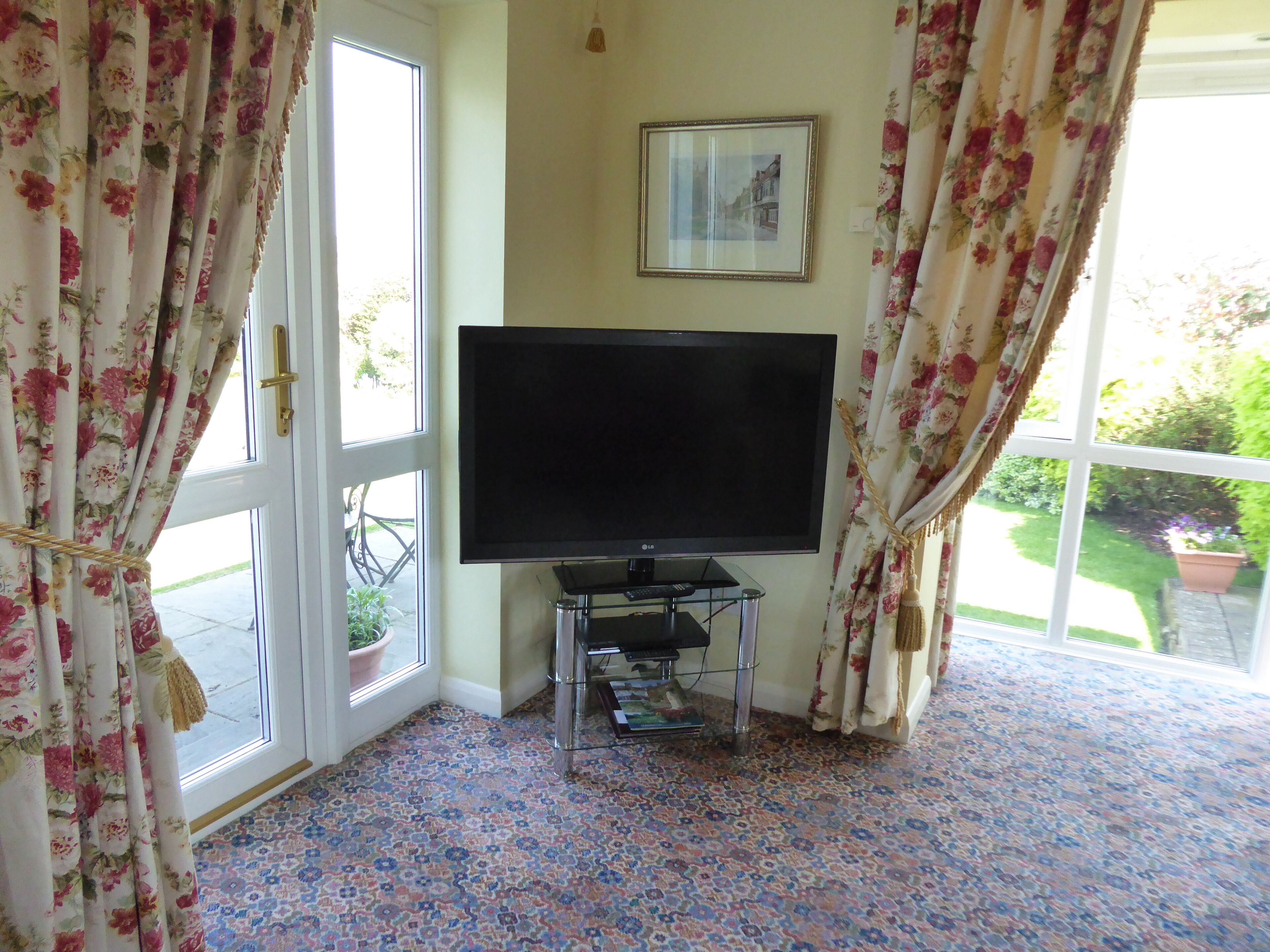 A room with a large flat-screen TV on a stand, floral curtains, a framed picture on the wall, and a potted plant by the window.