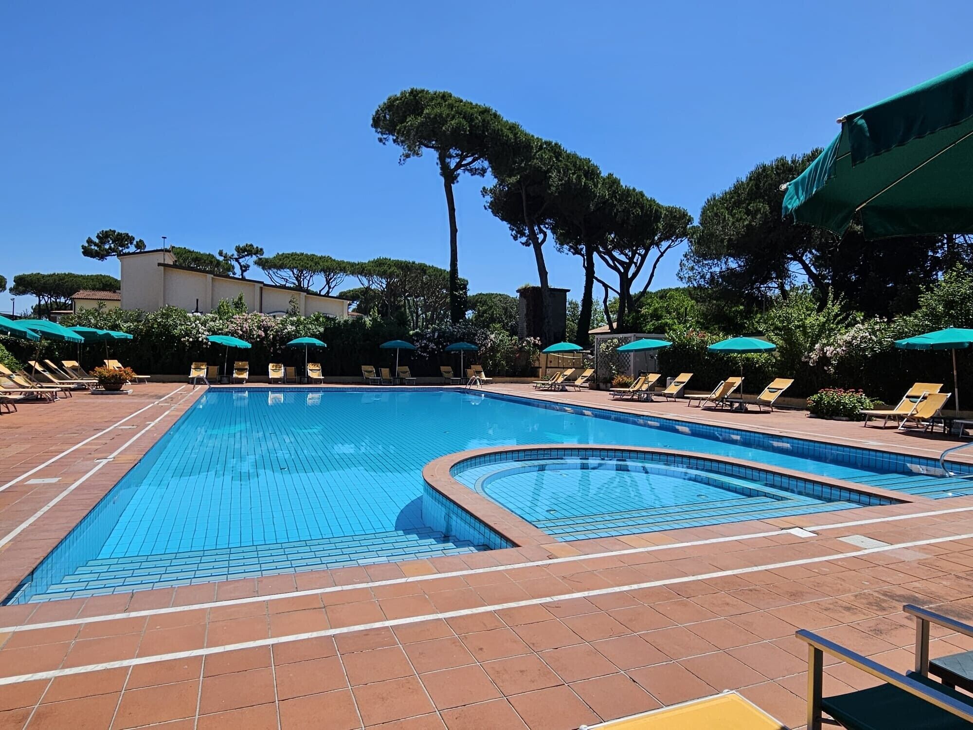A swimming pool with sun loungers and umbrellas, surrounded by trees and a building.