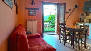 A cozy living room with a red sofa, a dining table with chairs, a radiator, and a view of greenery outside. - In Farmhouse independent apartment 2+2 people. Breathtaking view! (Urbino)