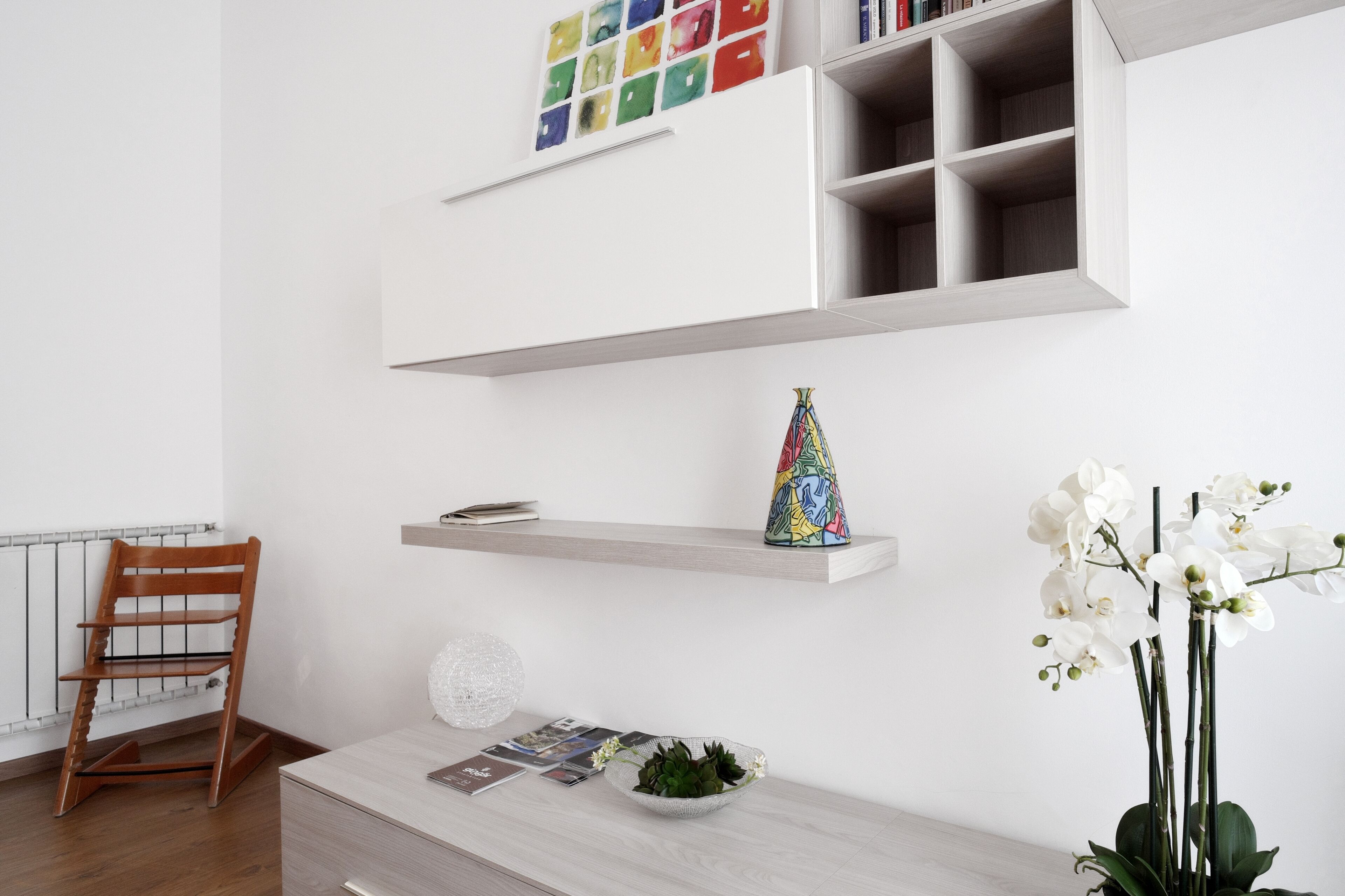 A modern room with a wooden chair, a white console table with decorative items, a bookshelf with colorful artwork, and a potted orchid plant.