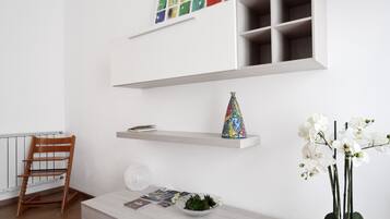A modern room with a wooden chair, a white console table with decorative items, a bookshelf with colorful artwork, and a potted orchid plant.