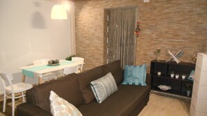 A living room with a brown sofa, a dining table with chairs, a stone wall, and a small shelf with decorative items.