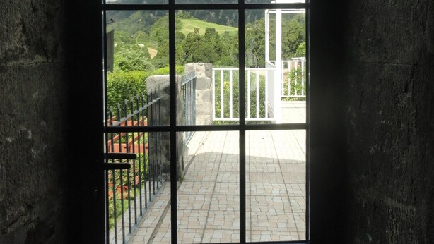 A window with a view of a green landscape, a white building, and a stone wall.