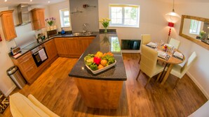 A modern kitchen with wooden cabinets, a central island with fresh produce, a dining area with a table set for two, and a large window providing natural light. - Heated indoor pool & spa, countryside holiday cottage near the beach & park (Hastings)