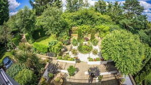 A lush garden with various plants and trees, a central circular area with seating, and a paved walkway. - Heated indoor pool & spa, countryside holiday cottage near the beach & park (Hastings)