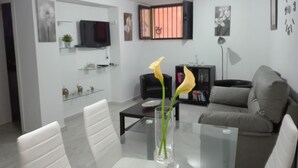 Una sala de estar moderna con una mesa de centro de vidrio, sillas blancas, un sofá gris y un televisor montado en la pared.