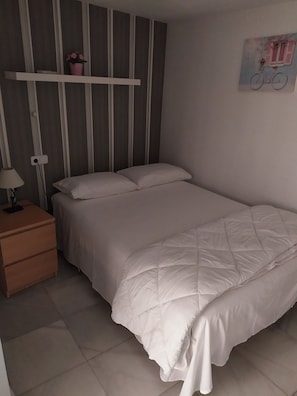 Una cama individual con un edredón blanco acolchado, una mesita de noche con una lámpara y una estantería con una planta en maceta.
