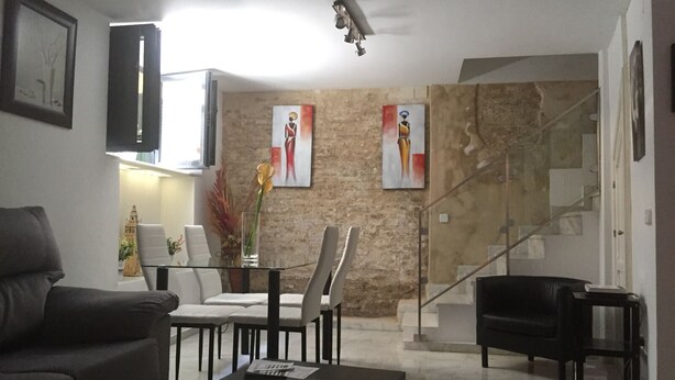 Una sala moderna con sofá, mesa de centro y zona de comedor con sillas. Hay dos cuadros en la pared, una escalera y una araña.