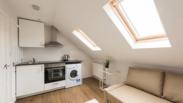 Une cuisine compacte équipée d’un lave-linge, d’une cuisinière et d’un four encastré. Le coin salon comprend un canapé et une petite table.