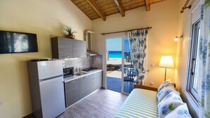 Una cocina compacta con refrigerador, estufa y pileta, un televisor de pantalla plana sobre la encimera, y una puerta corrediza de vidrio que da a un balcón con vista al mar.