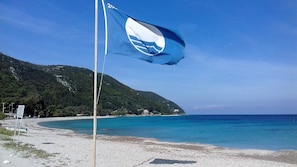 Um mastro com uma bandeira, uma praia e montanhas ao fundo.