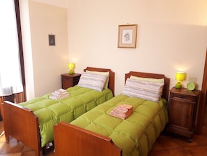 A double room with two beds, each with green bedding and wooden headboards. There are bedside tables with lamps and a framed picture on the wall.