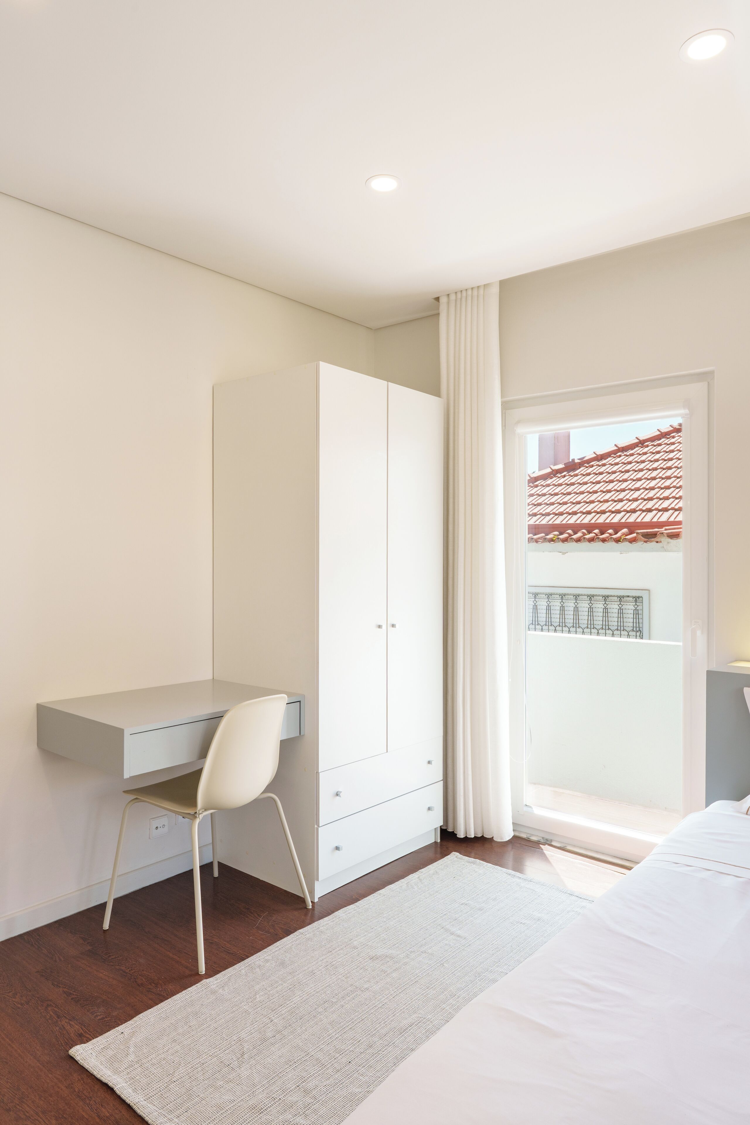A bedroom with a desk, chair, wardrobe, and a view of a balcony.