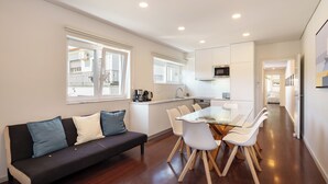 A modern kitchen with white cabinets, a dining table with white chairs, and a dark-colored sofa with blue cushions. - Apartment 4 bedrooms (2) - Porto (Porto)