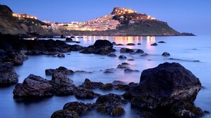 Un pueblo costero con una fortaleza imponente en una colina, costa rocosa y mar tranquilo.