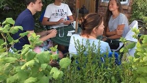 Een groep mensen zit aan een buitentafel onder een groene parasol, met een bakstenen huis en begroening op de achtergrond.
